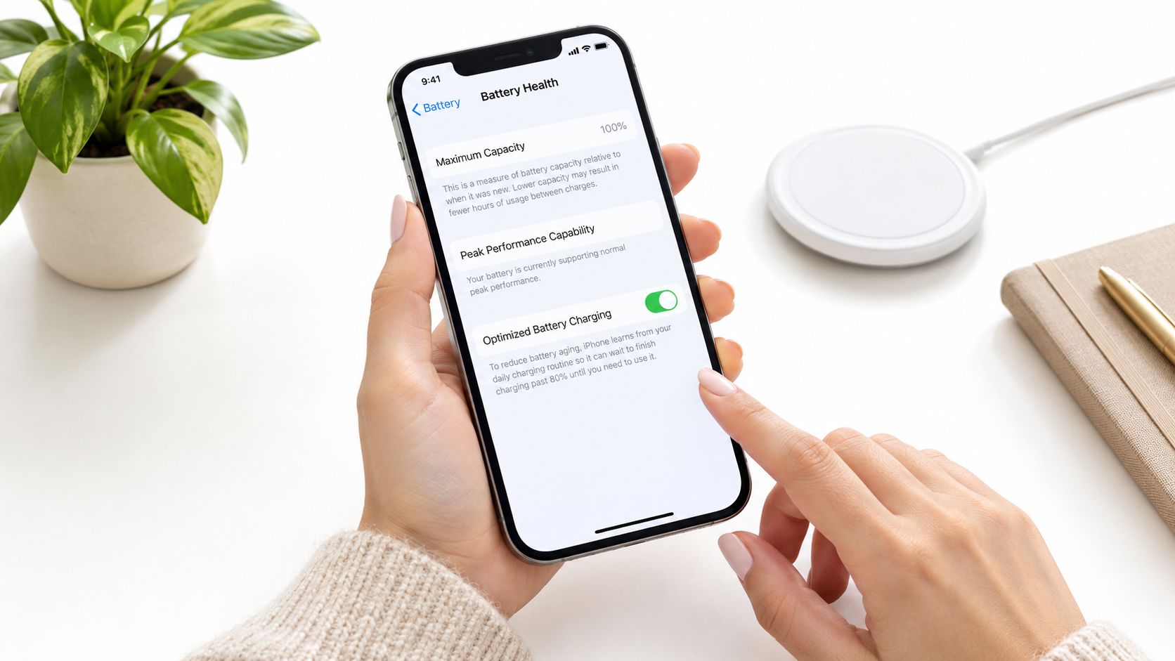A person checking their iPhone's battery health settings on a white desk with a wireless charger.